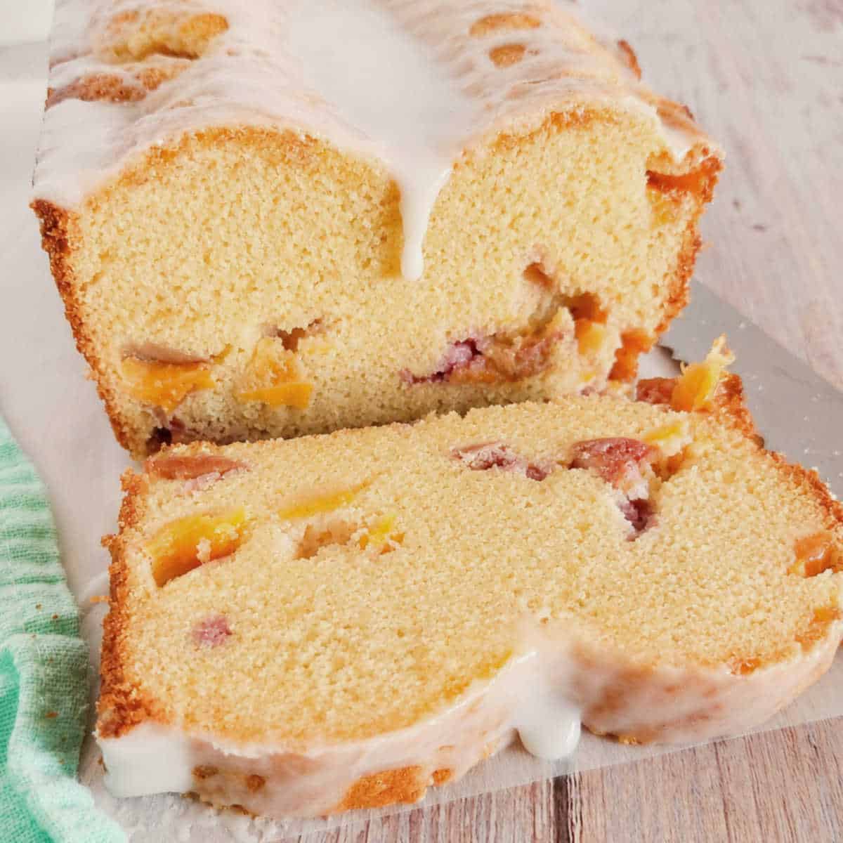 Iced peach loaf cake with two slices cut, showing chunks of peaches inside, on a light wooden surface next to a green striped cloth.