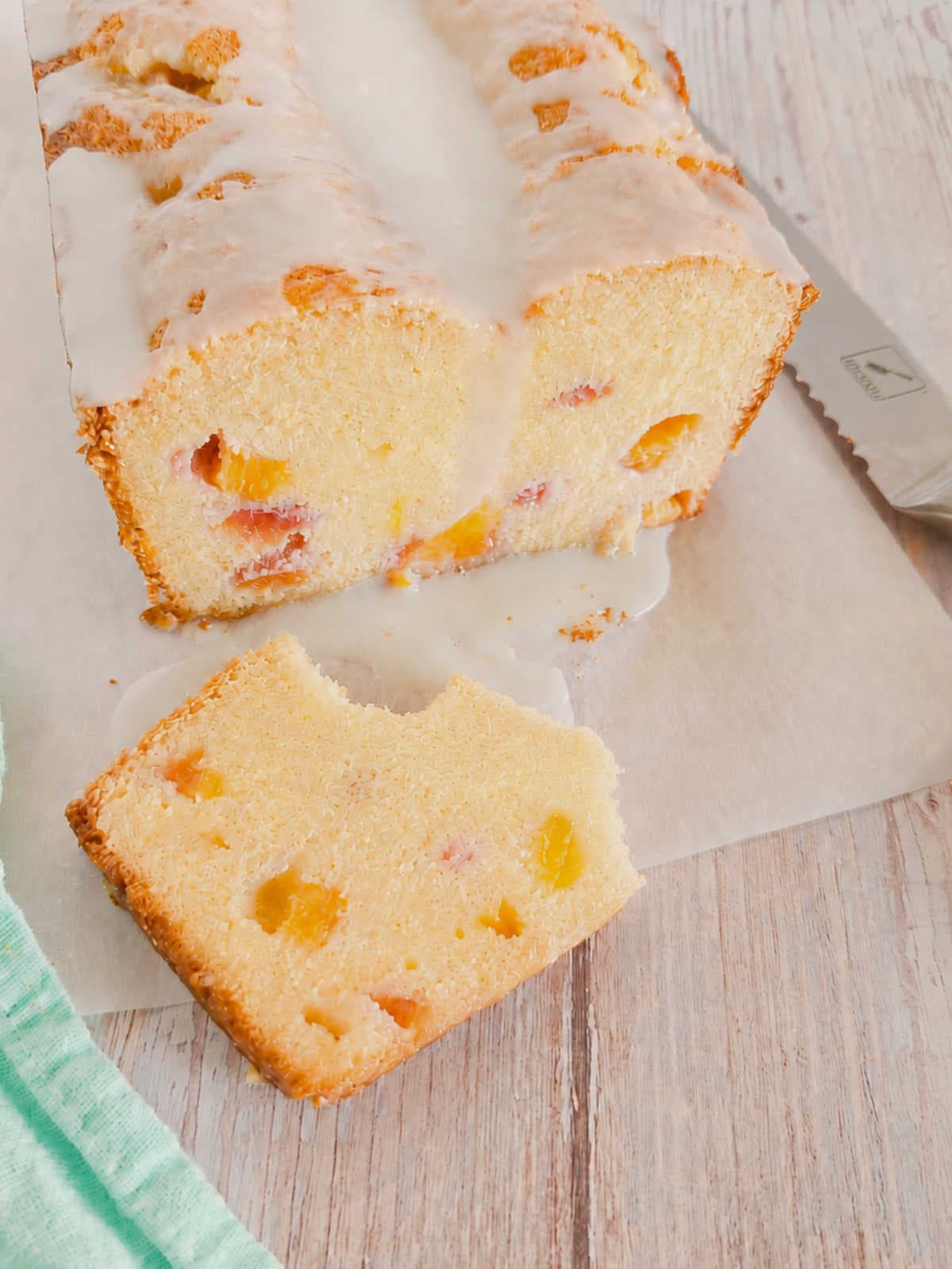 A loaf of glazed peach cake sits on parchment paper, with one slice cut and placed in front; a knife rests nearby.