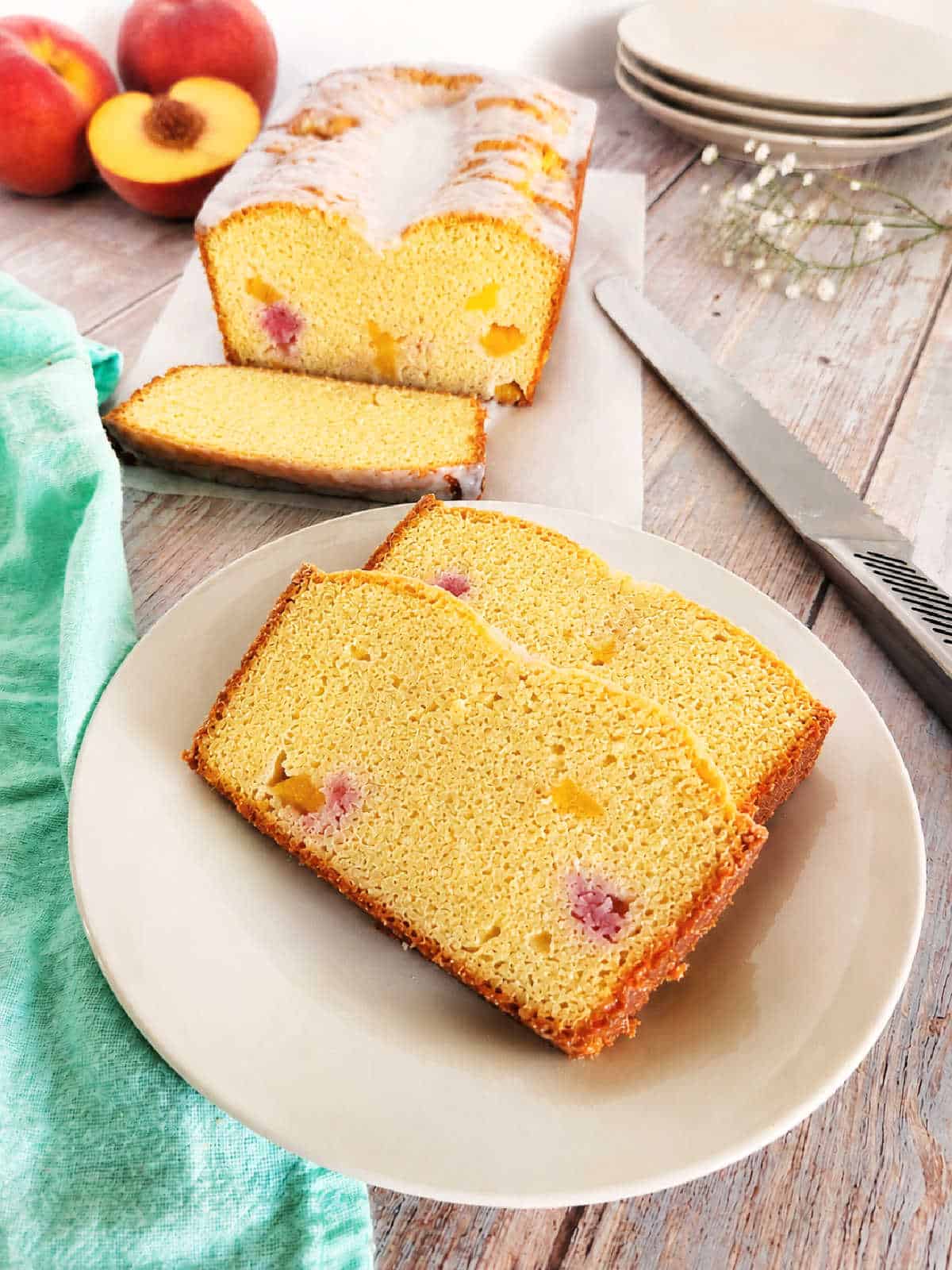 Two slices of peach bread are served on a white plate, with a loaf of the same bread and a knife on a cutting board in the background. Fresh peaches are also visible.