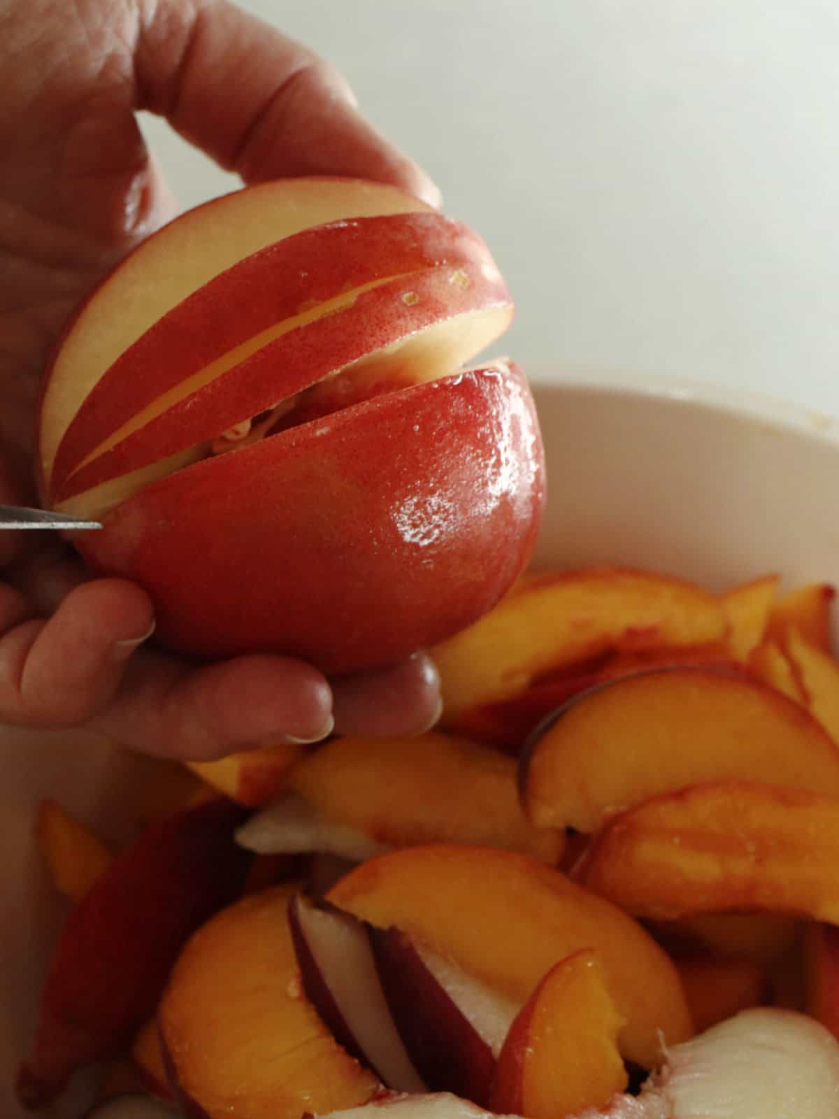 A hand slices a red nectarine above a bowl filled with nectarine wedges.
