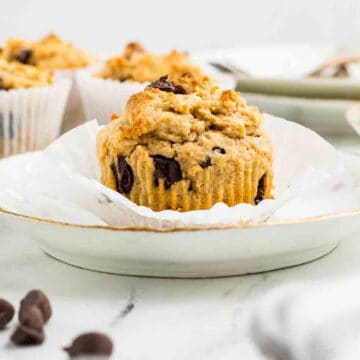 A peanut butter and chocolate chip muffin sits on a white plate with its paper wrapper partially peeled back; other muffins are in the background.