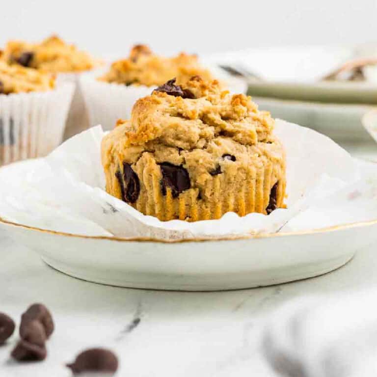 A peanut butter and chocolate chip muffin sits on a white plate with its paper wrapper partially peeled back; other muffins are in the background.