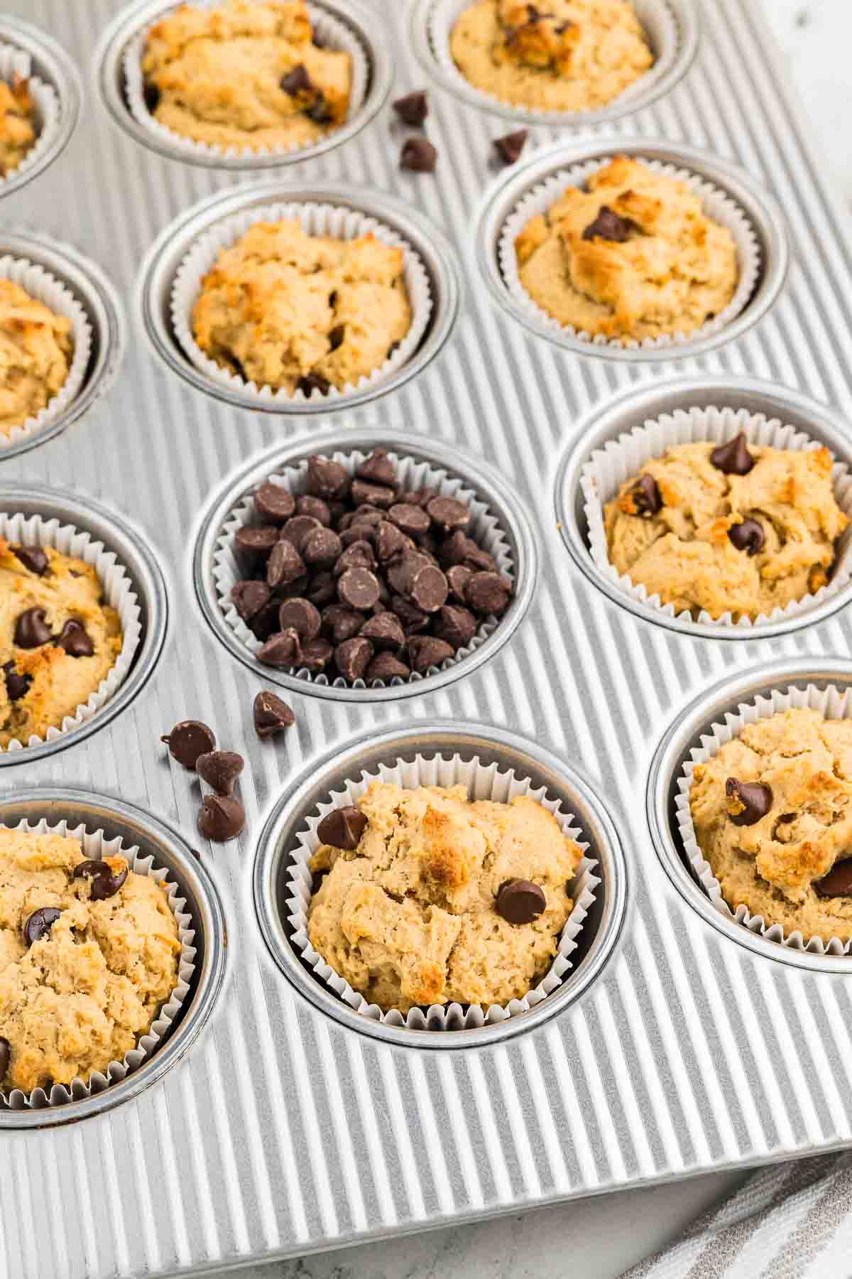 A muffin tin holds several baked chocolate chip muffins in paper liners, with one cup filled with chocolate chips instead of a muffin.