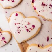 Heart-shaped sugar cookies with white icing and dried rose petal toppings on parchment paper and a wooden board.