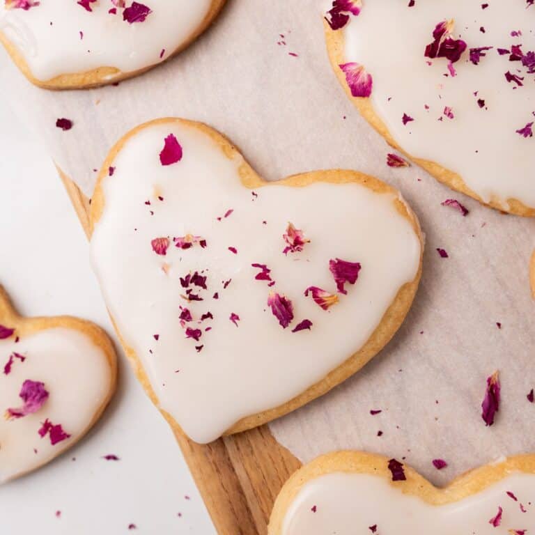 Heart-shaped sugar cookies with white icing and dried rose petal toppings on parchment paper and a wooden board.