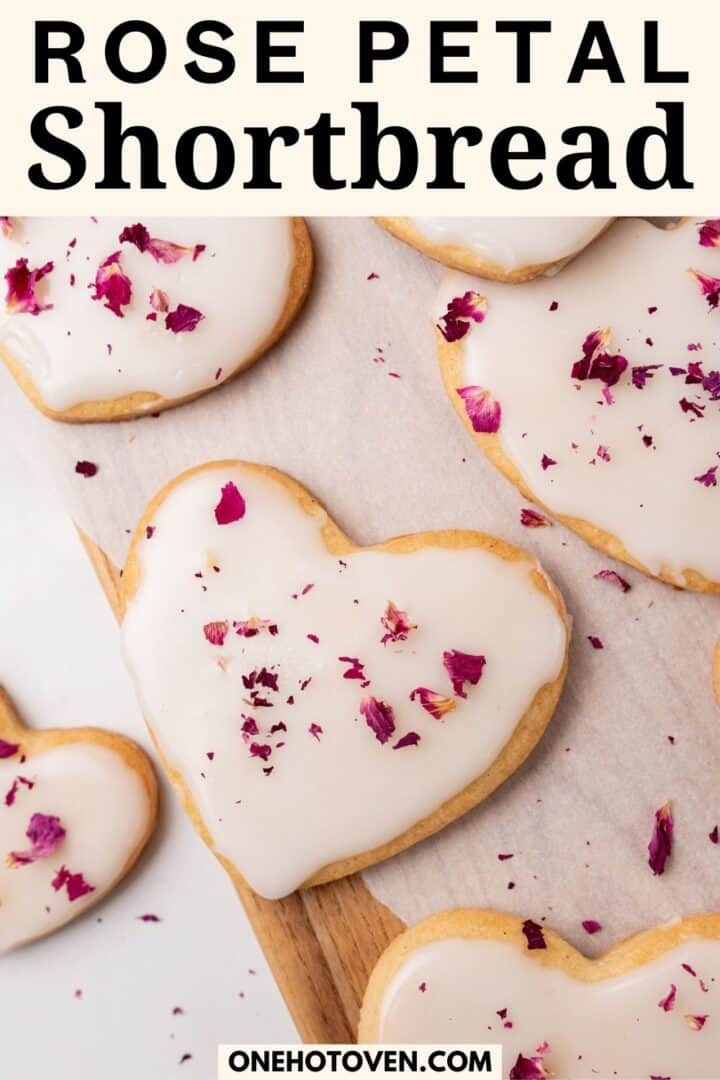 Heart-shaped shortbread cookies with white icing and sprinkled rose petals on parchment paper.