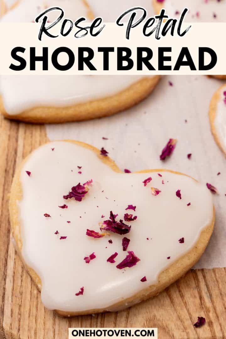 Heart-shaped shortbread cookie with white icing and rose petal pieces on top, placed on a wooden surface with parchment paper.