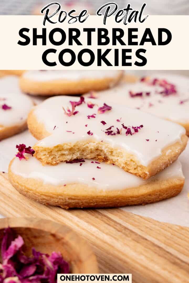 Iced shortbread cookies topped with rose petals are arranged on a wooden board, with one cookie showing a bite taken out.