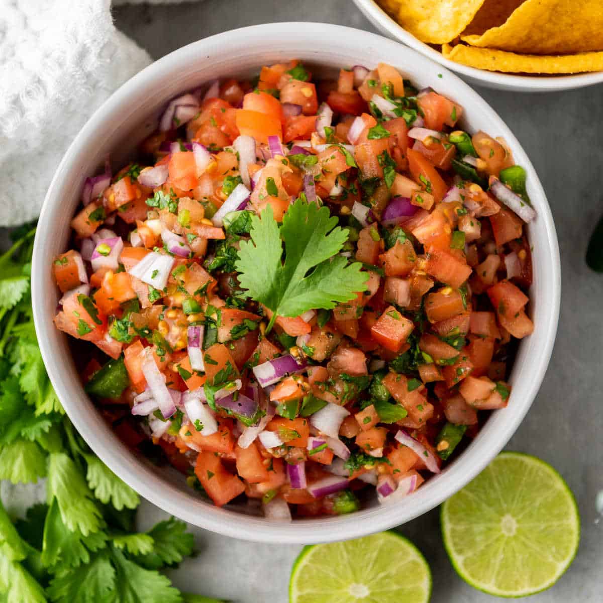 A bowl of pico de gallo made with diced tomatoes, red onions, cilantro, and jalapeños, garnished with a cilantro leaf, surrounded by lime halves and tortilla chips.