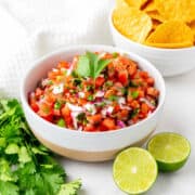 A bowl of fresh salsa with diced tomatoes, onions, cilantro, and peppers, garnished with a cilantro sprig. Sliced lime, cilantro leaves, and a bowl of tortilla chips are nearby.