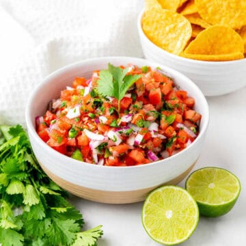 A bowl of fresh salsa with diced tomatoes, onions, cilantro, and peppers, garnished with a cilantro sprig. Sliced lime, cilantro leaves, and a bowl of tortilla chips are nearby.