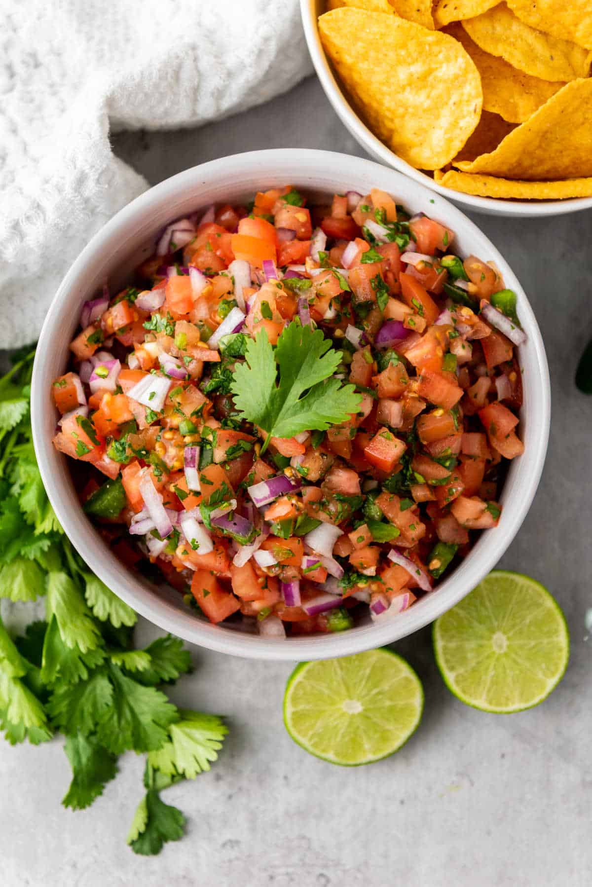 A bowl of fresh pico de gallo garnished with cilantro, surrounded by lime halves, cilantro leaves, and a bowl of tortilla chips.