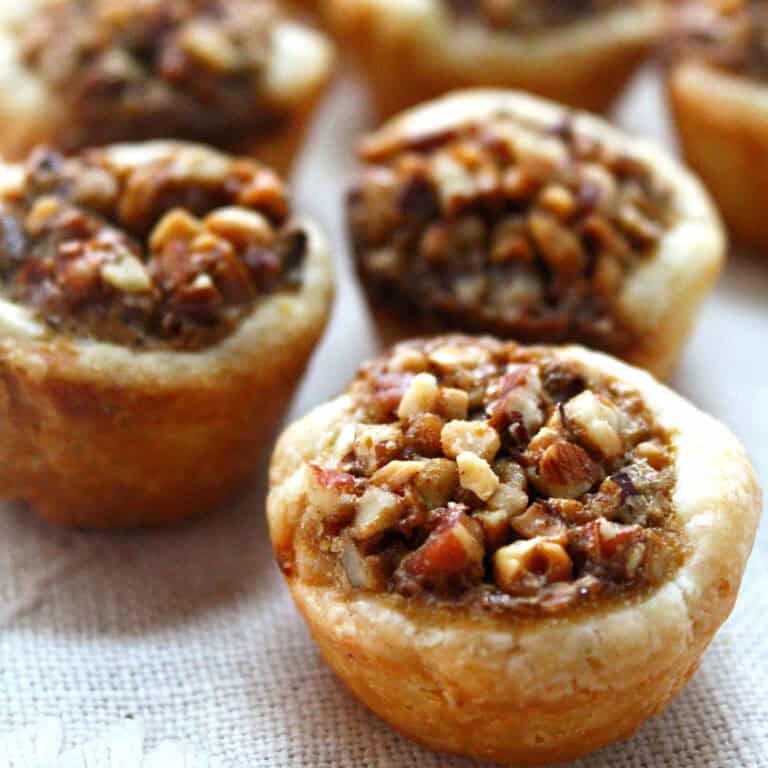 Close-up of several mini pecan pies with golden crusts and chopped pecan topping, arranged on a light-colored cloth.