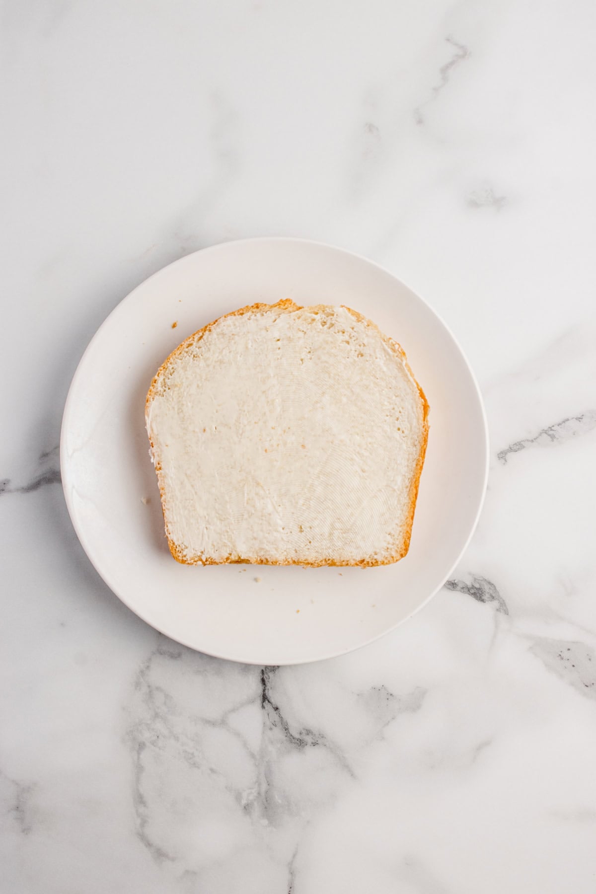 A single slice of white bread with a layer of butter on a white plate, placed on a marble surface.