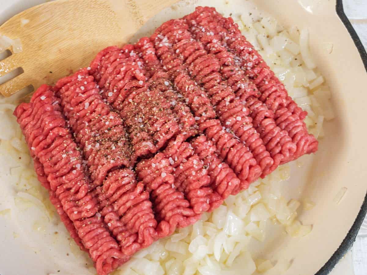 Raw ground beef with salt and pepper sits on sautéed onions in a skillet, with a wooden spatula nearby.