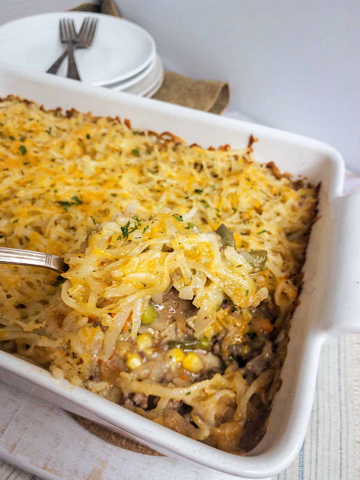 A baking dish filled with a hambuger, shredded potato casserole topped with shredded cheese and hash browns, with a serving spoon lifting a portion. Plates, forks, and a napkin are in the background.
