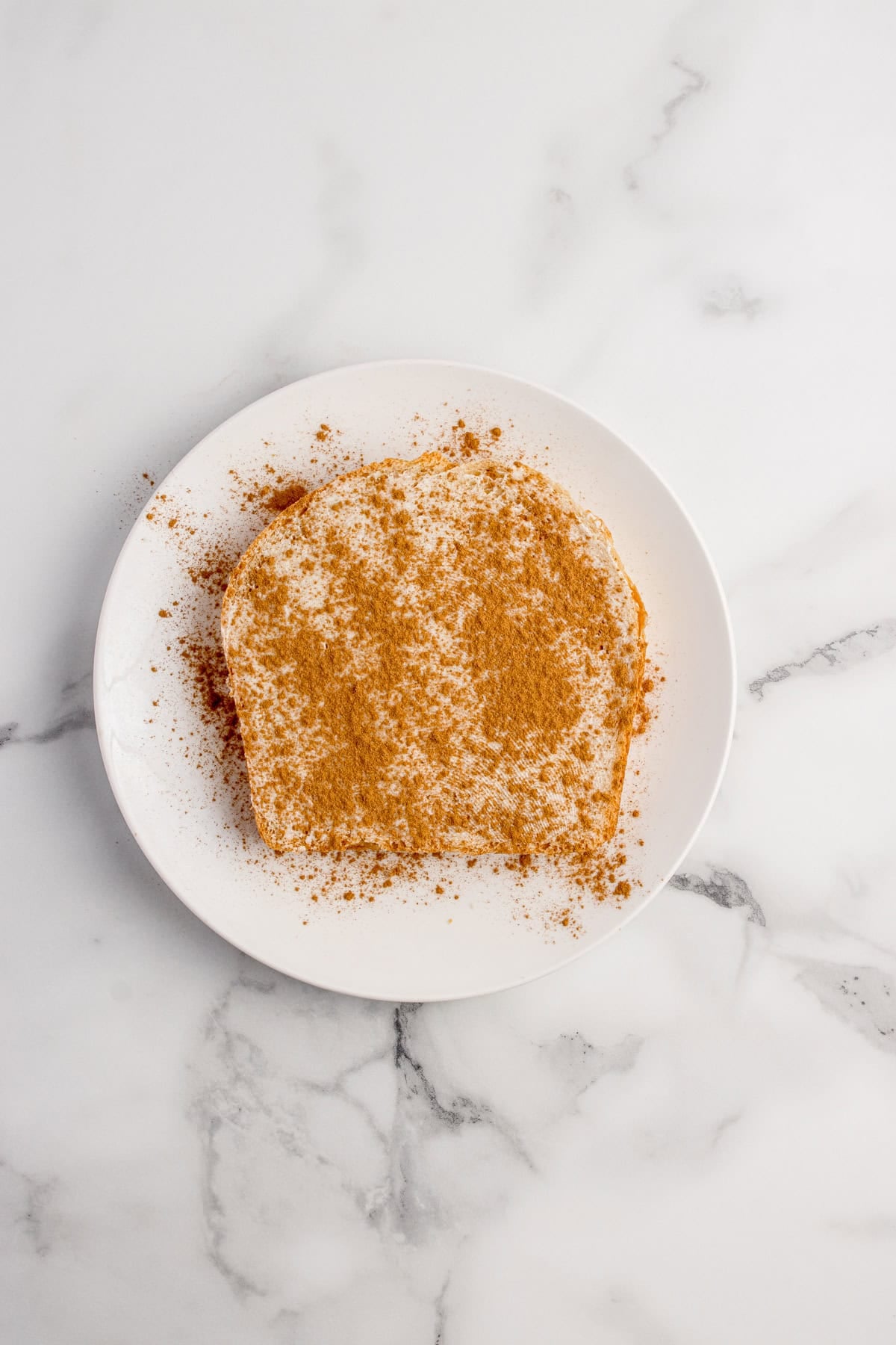 A slice of bread covered with cinnamon and sugar sits on a white plate atop a marble surface.