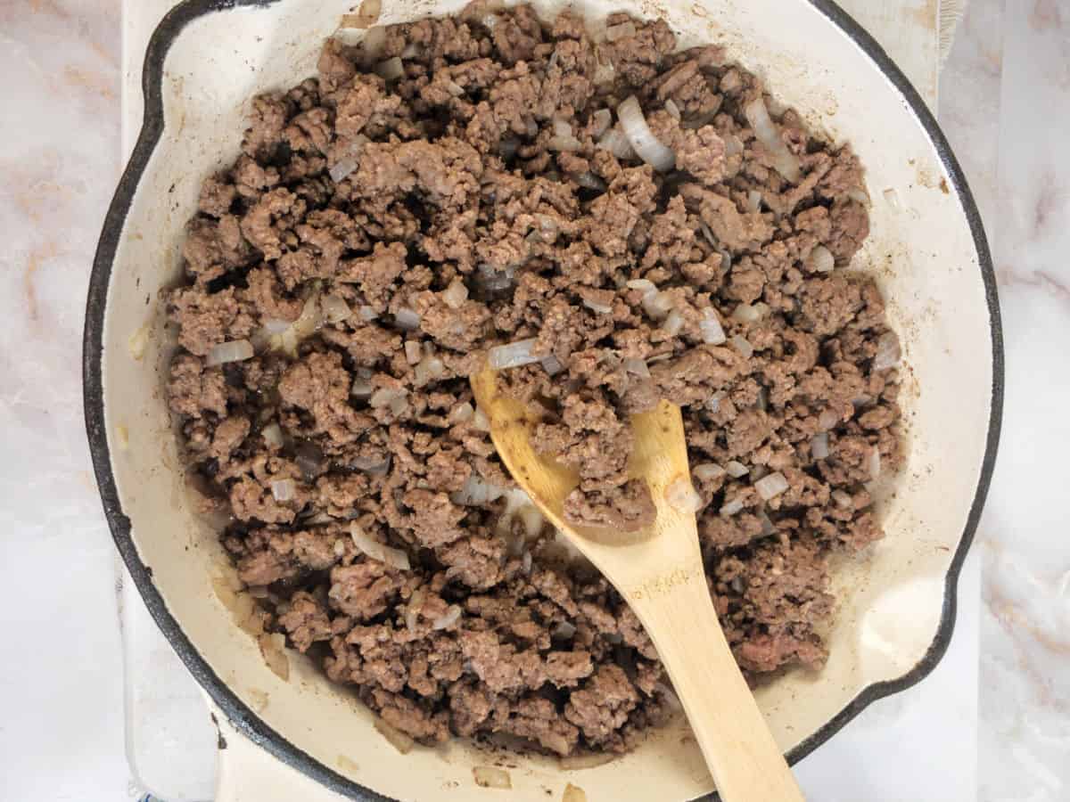 Cooked ground beef and chopped onions in a white skillet, being stirred with a wooden spoon.