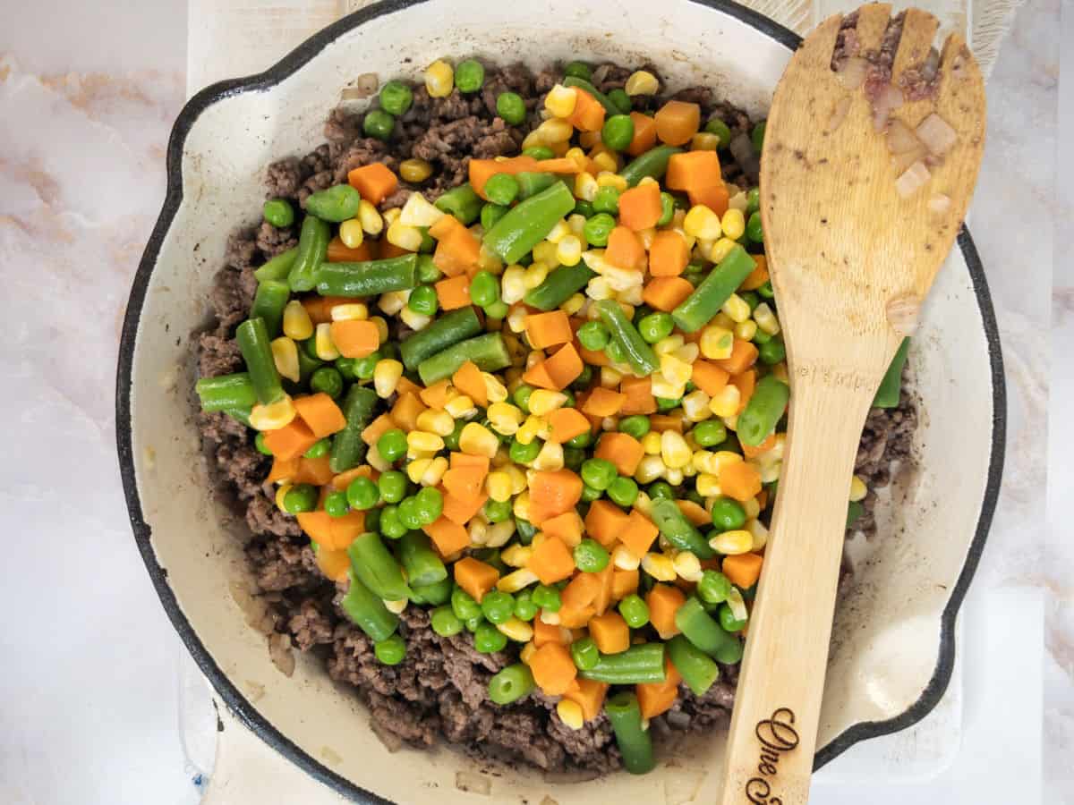 A skillet filled with cooked ground beef topped with a colorful mix of green beans, carrots, corn, and peas. A wooden spoon with chopped onions rests on the side.