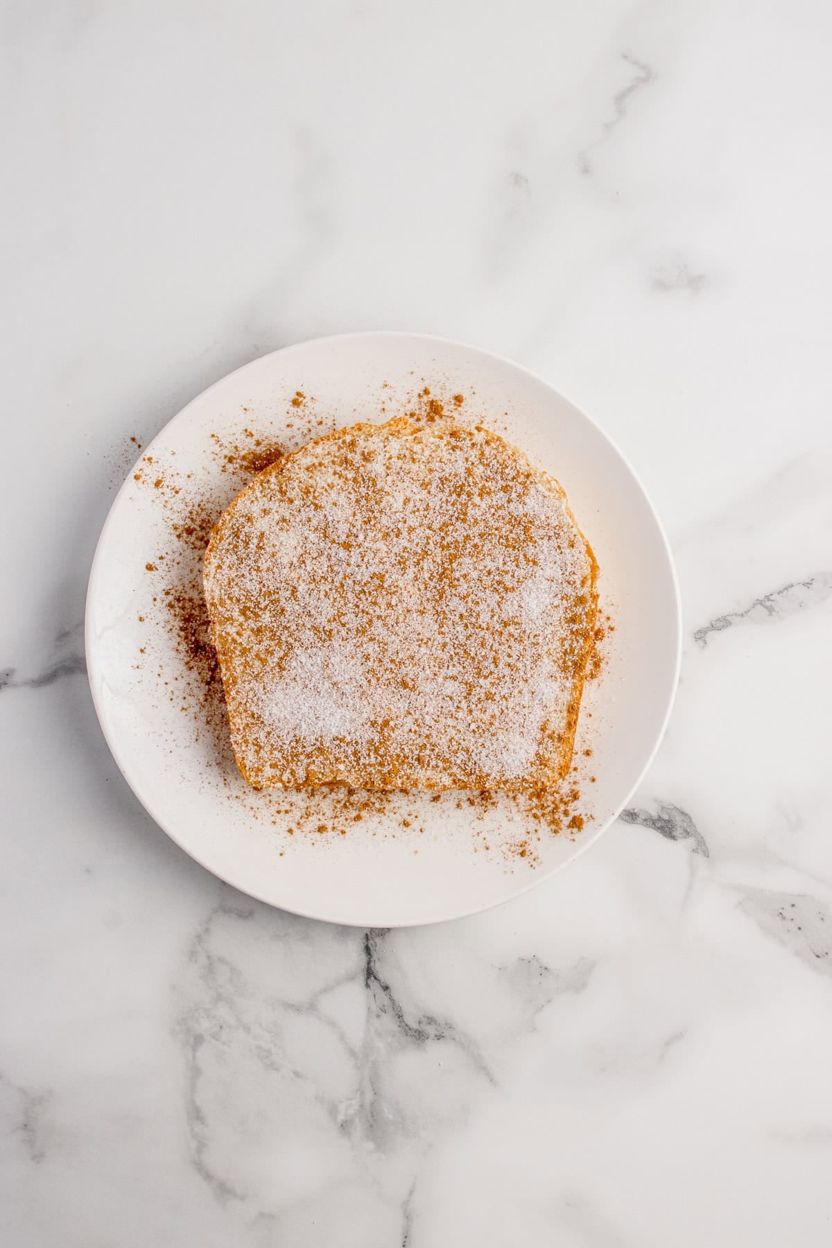 A slice of bread covered with a layer of sugar and cinnamon sits on a white plate atop a marble surface.
