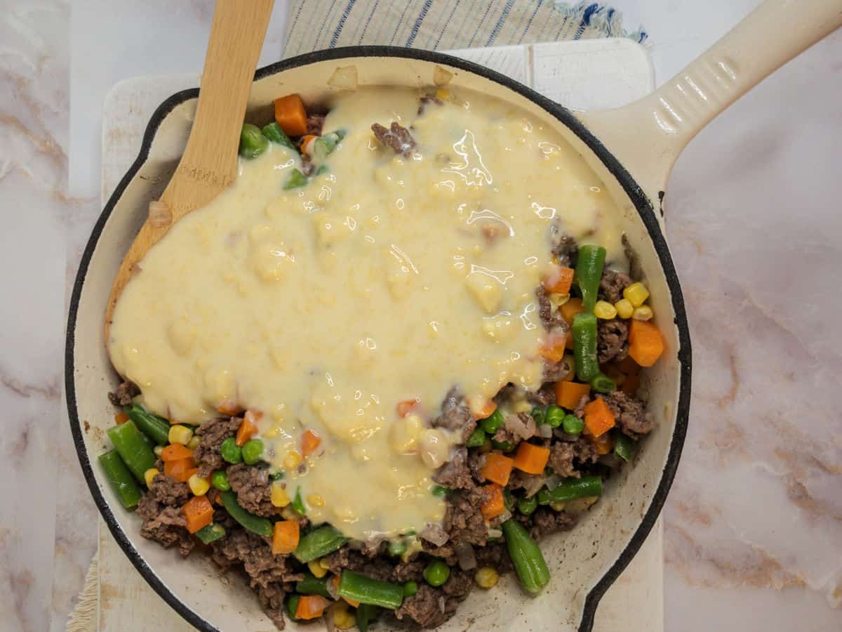 A skillet contains ground beef, mixed vegetables, and cream soup being stirred with a wooden spoon.