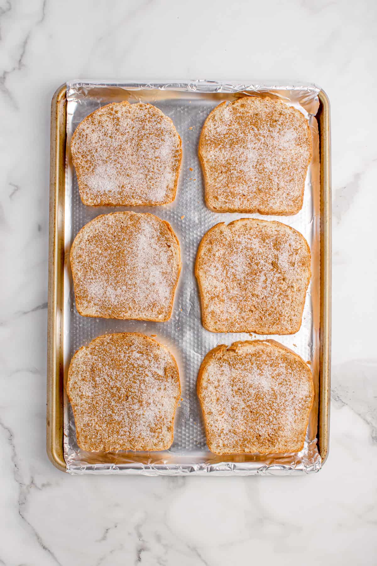 A baking sheet lined with foil holds six slices of white bread topped with a cinnamon-sugar mixture, arranged in two rows on a marble surface for broiled cinnamon toast.