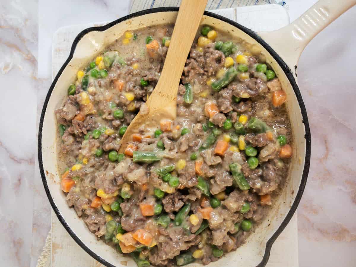 A skillet filled with a mixture of ground beef, green beans, peas, carrots, and corn in a creamy sauce, with a wooden spoon resting inside.