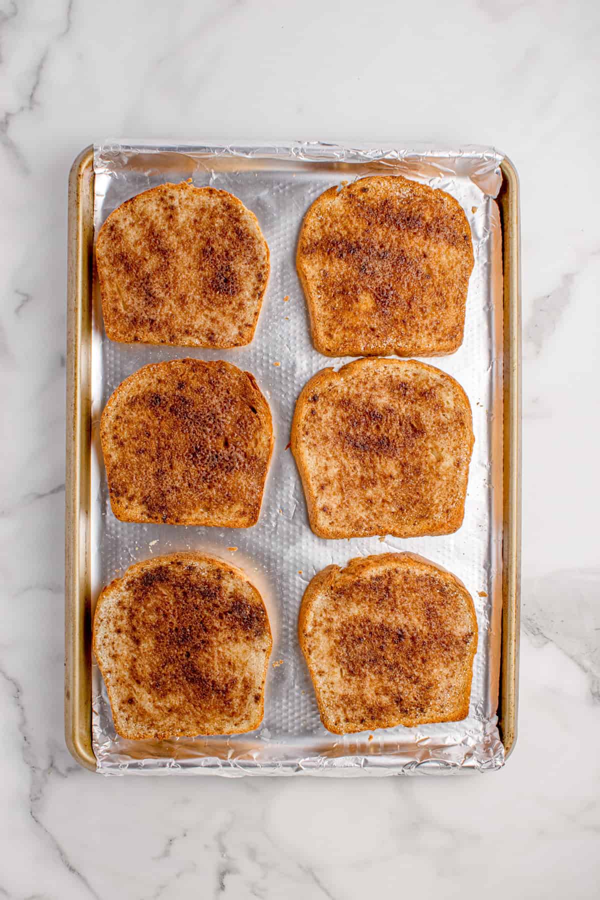Six slices of bread topped with cinnamon and sugar are arranged on a foil-lined baking sheet, set on a marble countertop.