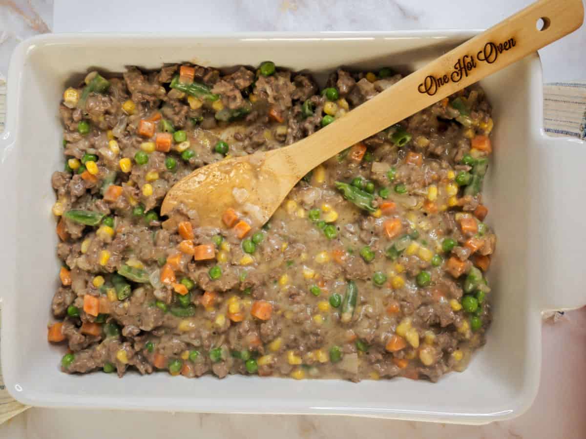 A baking dish filled with mixed vegetables and ground beef in a creamy sauce, being stirred with a wooden spoon labeled “One Hot Oven.”.