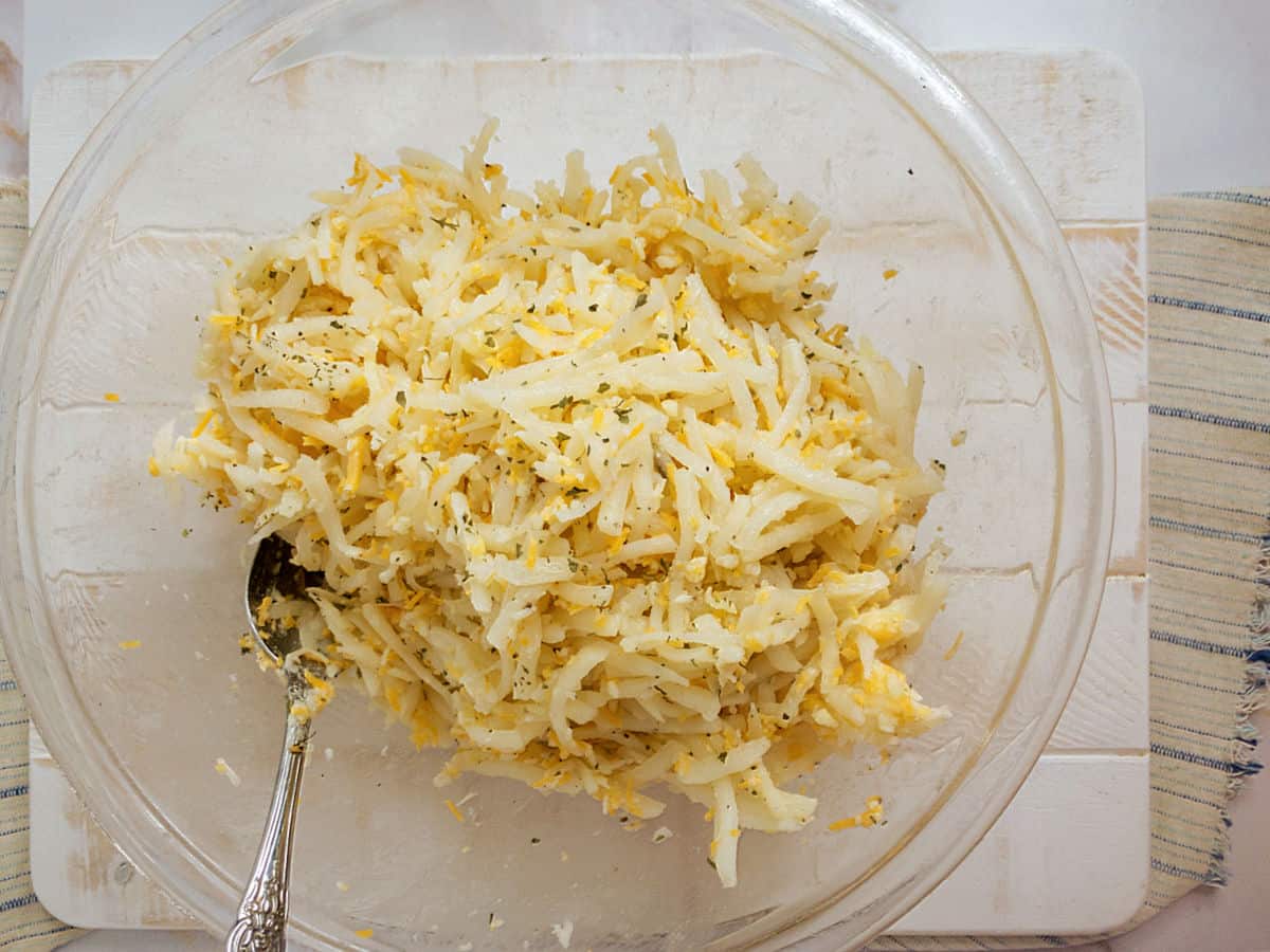 A clear bowl filled with shredded potatoes mixed with cheese and herbs; a spoon rests in the bowl, which sits on a white surface next to a striped cloth.