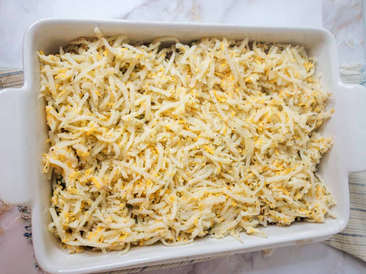 Rectangular white baking dish filled with an even layer of shredded cheese and potatoes, prepared for baking.