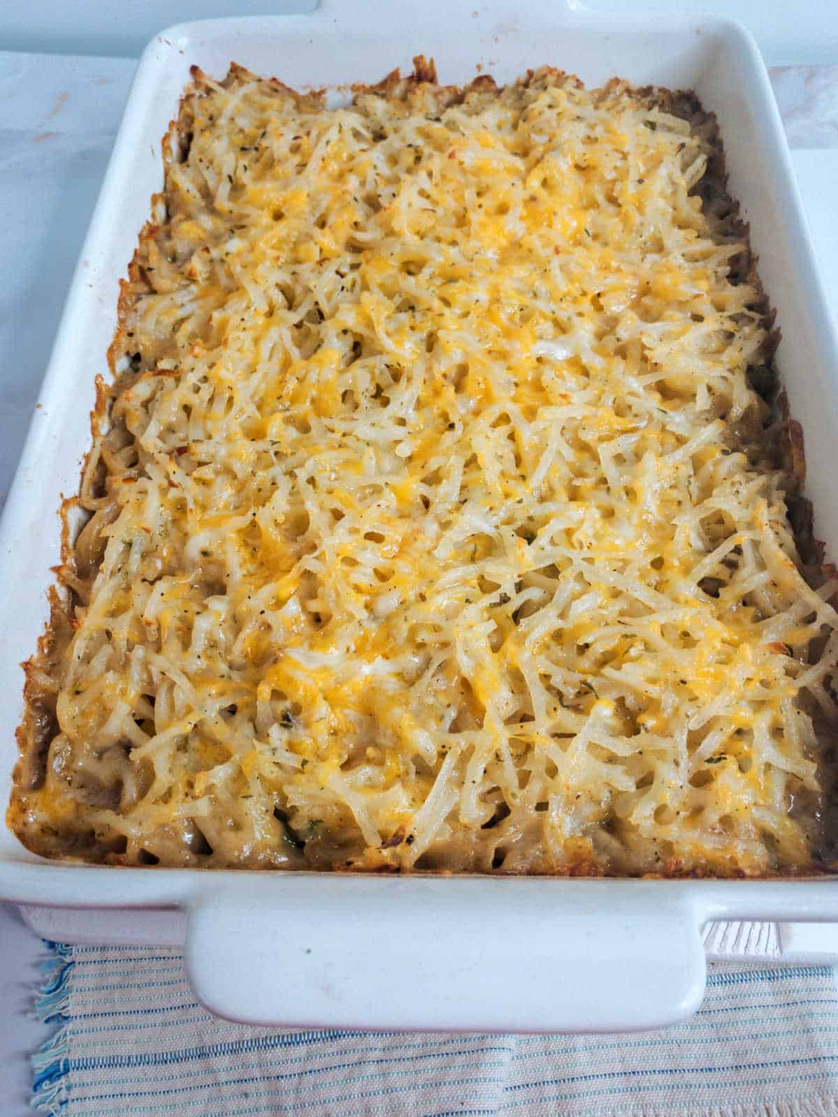 A rectangular white baking dish filled with a baked casserole topped with melted shredded cheese and hash browns, placed on a light blue striped cloth.