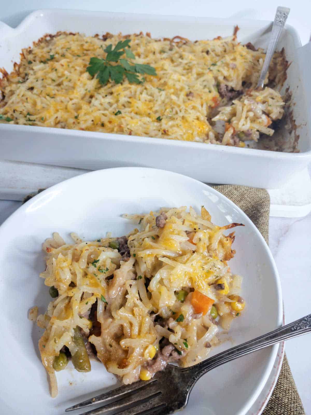 A serving of cheesy hamburger casserole with vegetables and ground meat on a white plate, fork beside it; baking dish with more casserole in the background.