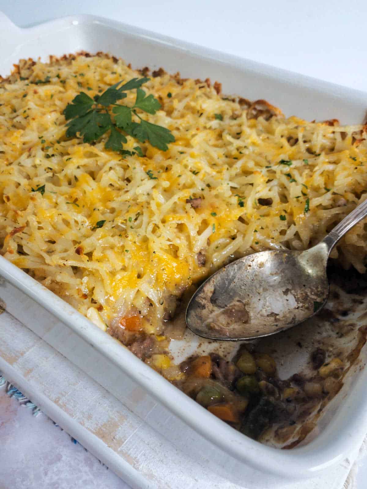 A hambuger casserole topped with shredded cheese and hash browns, partially served with a spoon in a white baking dish, garnished with a sprig of parsley.