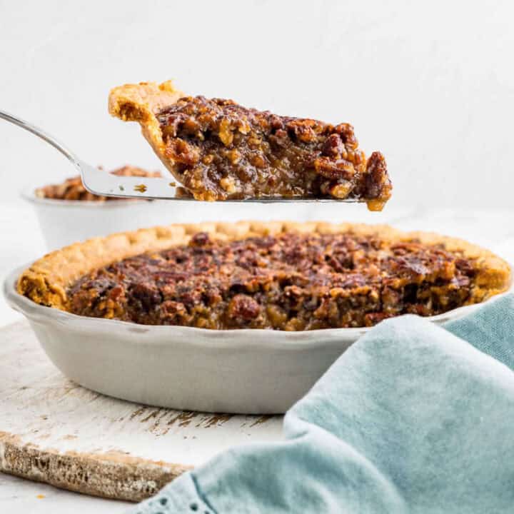 A slice of pecan pie is being lifted from a whole pie in a ceramic dish, with a light blue cloth in the foreground.