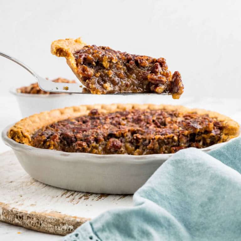 A slice of pecan pie is being lifted from a whole pie in a ceramic dish, with a light blue cloth in the foreground.
