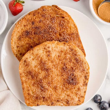 Two slices of toasted bread coated with cinnamon sugar on a white plate, surrounded by bowls of ingredients and fresh berries.