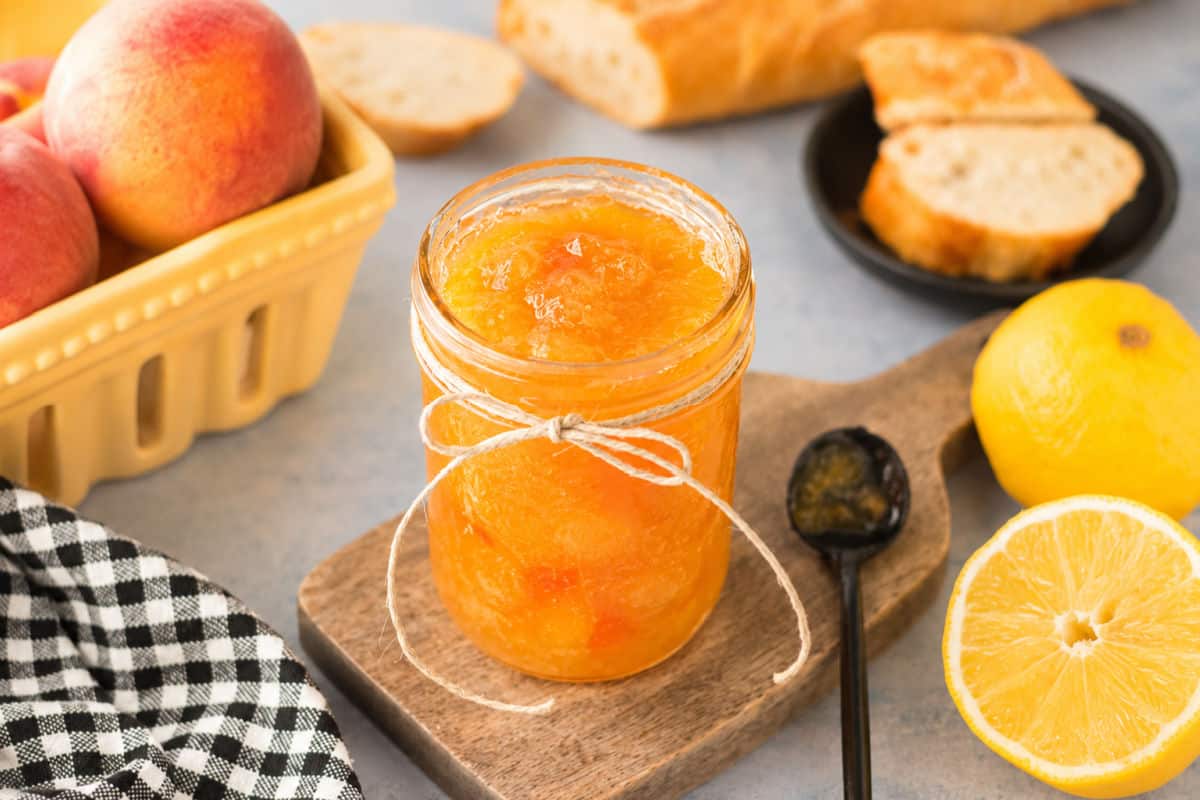 A jar of peach jam with a tied string sits on a wooden board beside a black spoon, lemon halves, sliced bread, fresh peaches, and a checked cloth.