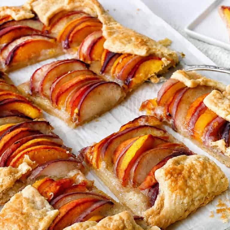 A sliced rustic peach tart with a flaky crust, topped with neatly arranged peach slices, sits on parchment paper.