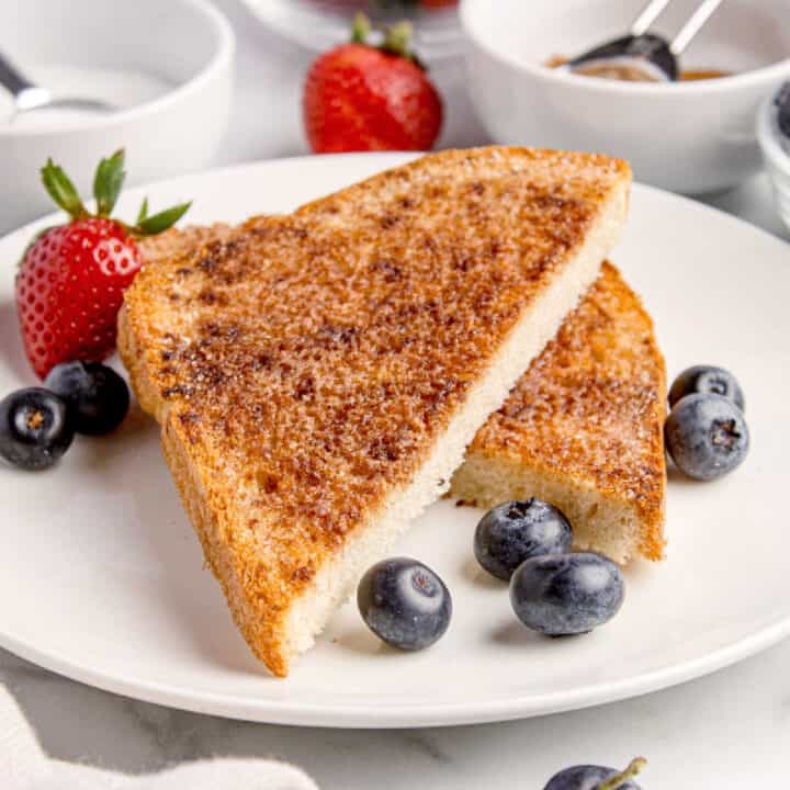 Two slices of broiled cinnamon toast on a white plate, garnished with fresh strawberries and blueberries.
