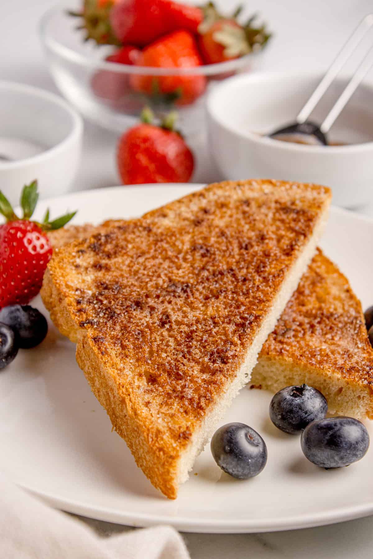 Two slices of cinnamon toast on a white plate with blueberries and a strawberry, with bowls of strawberries and sauces in the background.