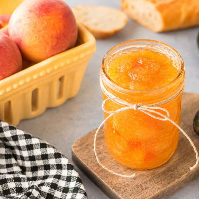 A jar of peach jam with a twine bow sits on a small wooden board, next to fresh peaches, sliced bread, and a black-and-white checkered cloth.