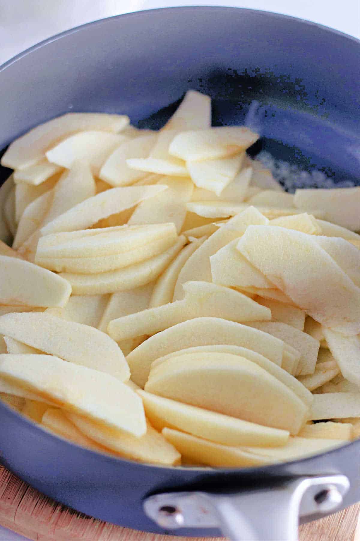 Thinly sliced apples Granny Smith apples in a blue skillet sitting on a wood board.