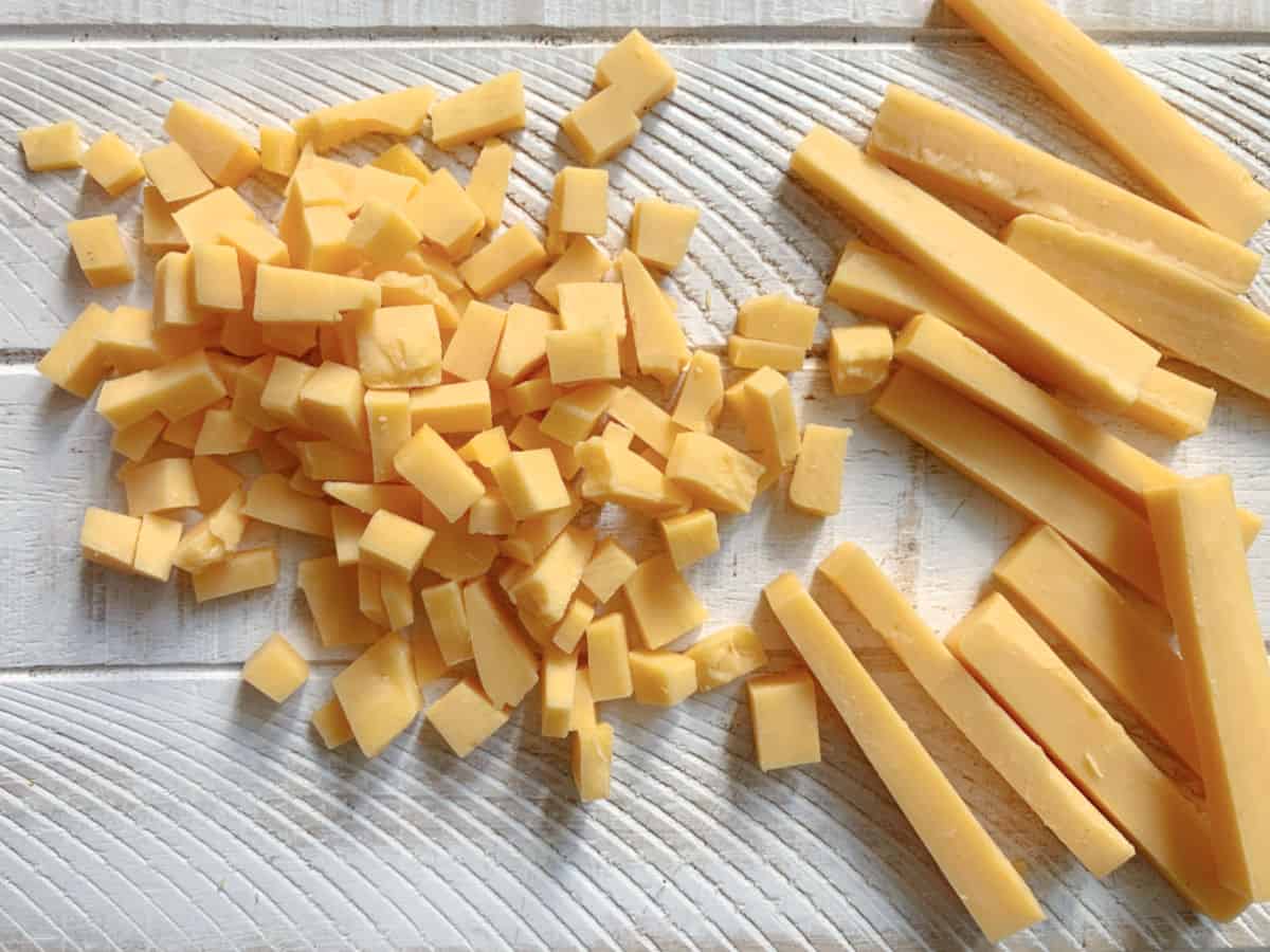 Cubed and sliced cheddar cheese arranged on a textured white wooden surface.