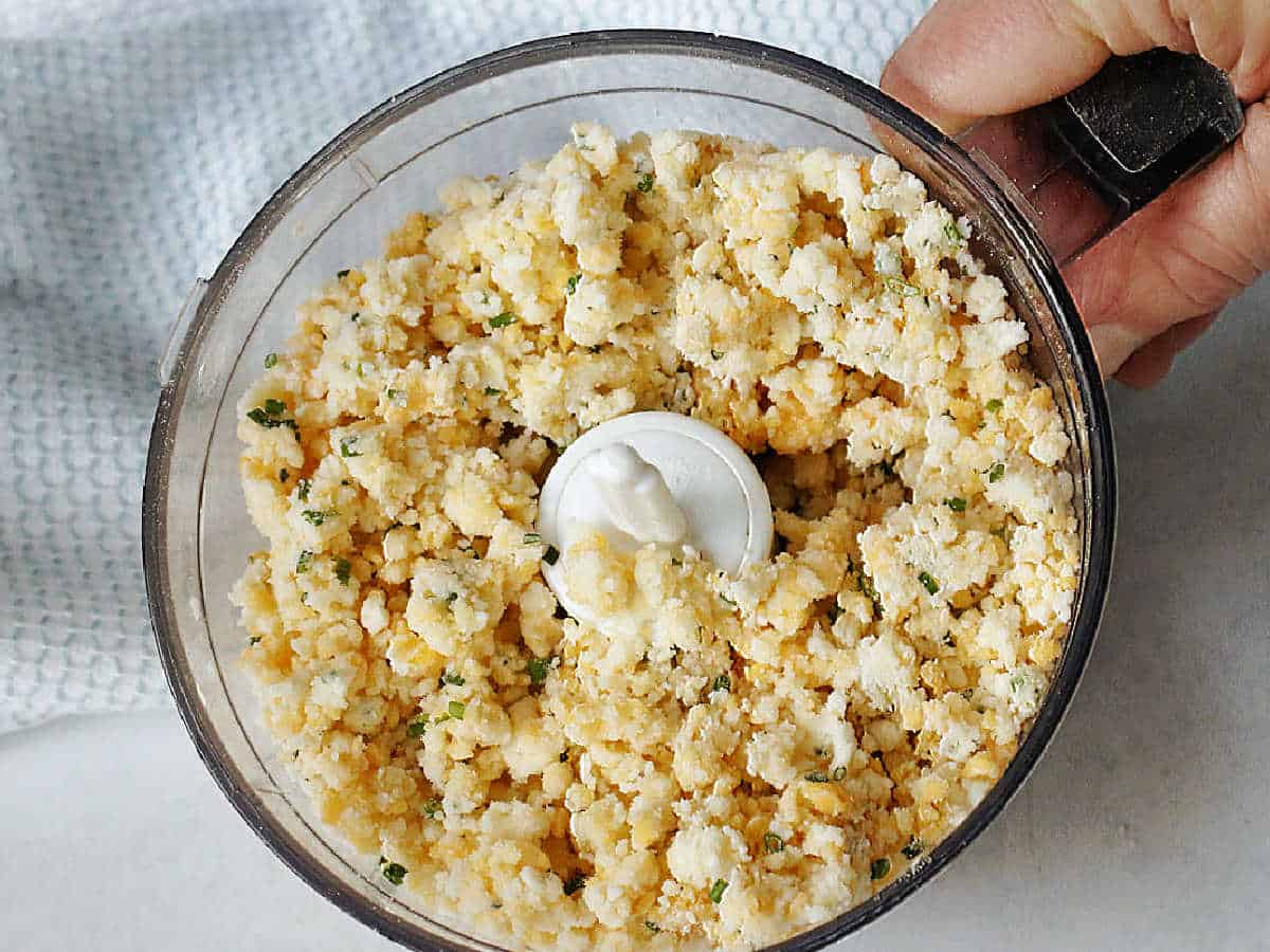 A food processor bowl filled with a crumbly cheese and herb mixture, shown from above with a hand holding the handle.