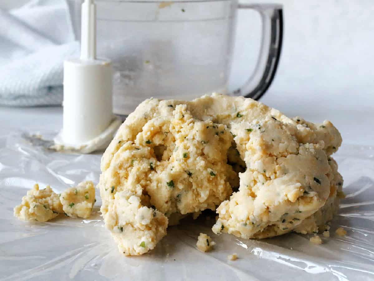 A ball of herb-flecked cracker dough rests on plastic wrap next to a food processor with a white blade attachment.