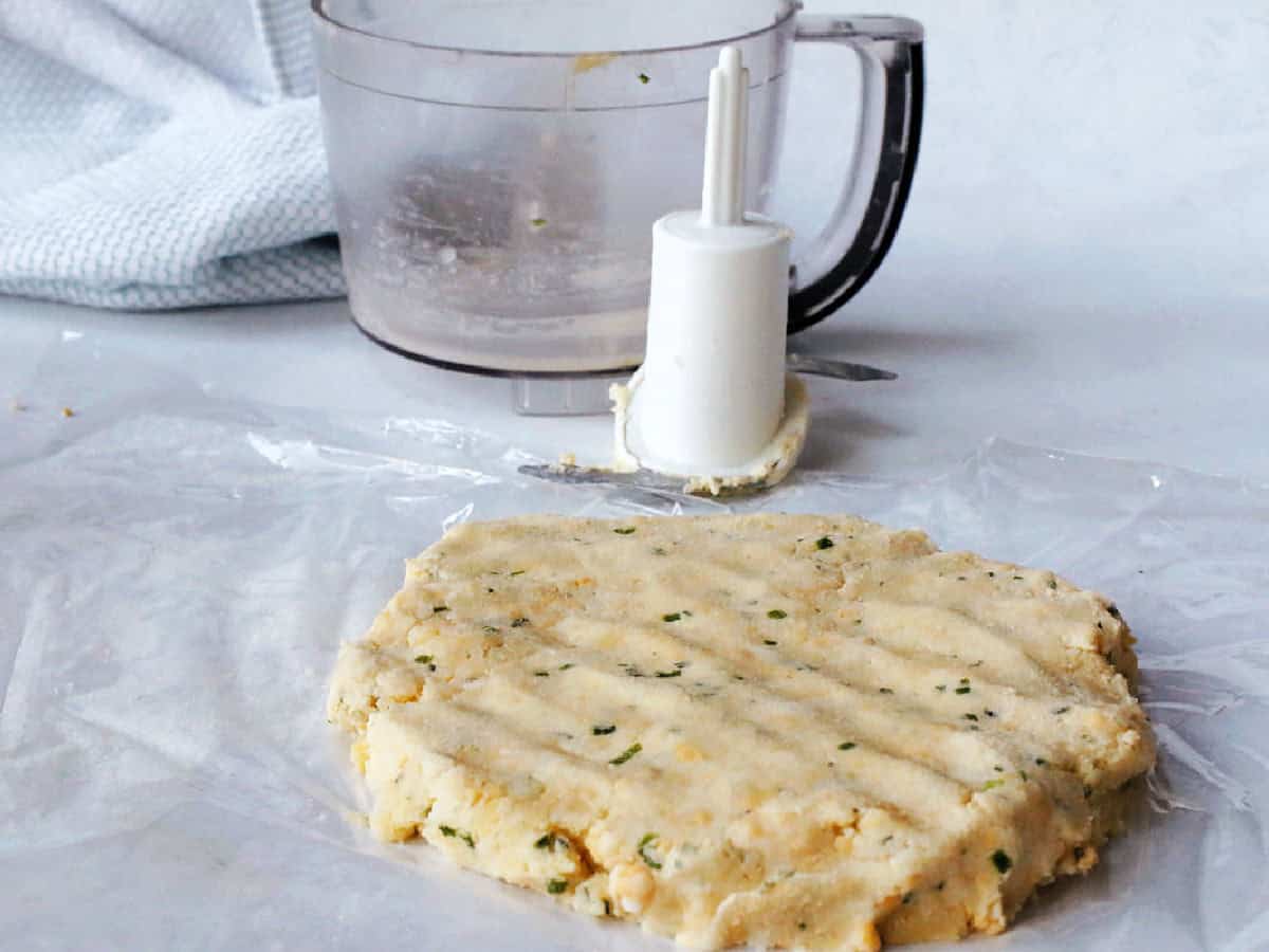 A flattened disc of cheddar cheese cracker dough with herbs sits on wax paper near a food processor with a towel in the background.