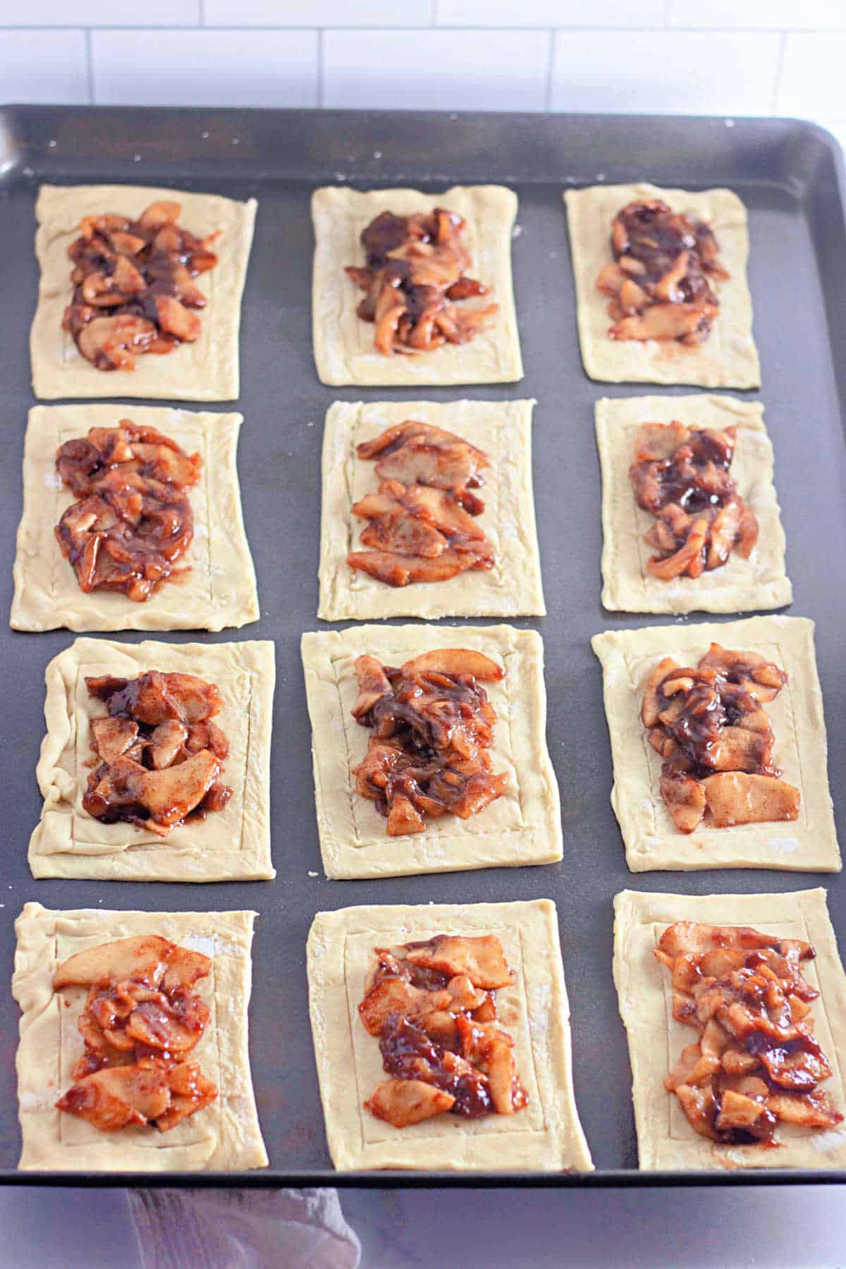 A baking sheet with twelve rectangles of pastry dough topped with cinnamon apple filling.