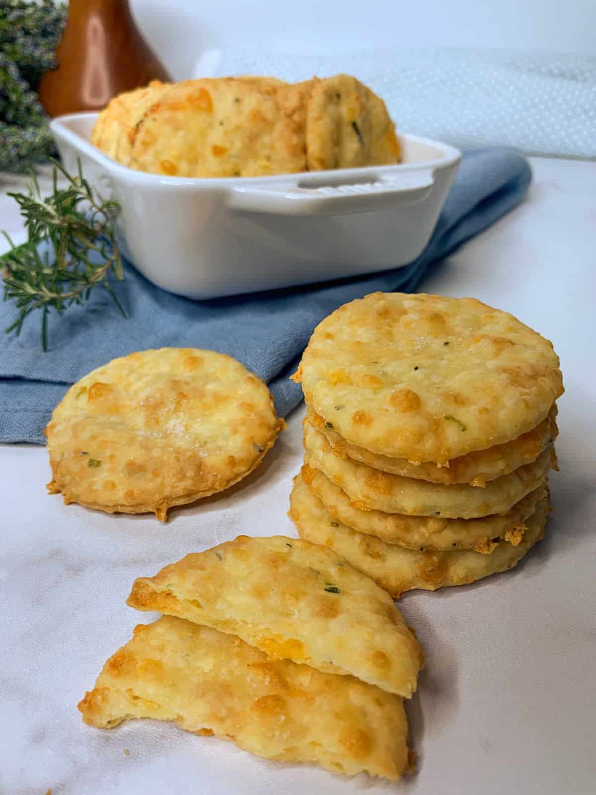 A stack of round, golden cheese crackers sits on a white surface, with more crackers in a white dish and a blue napkin in the background.