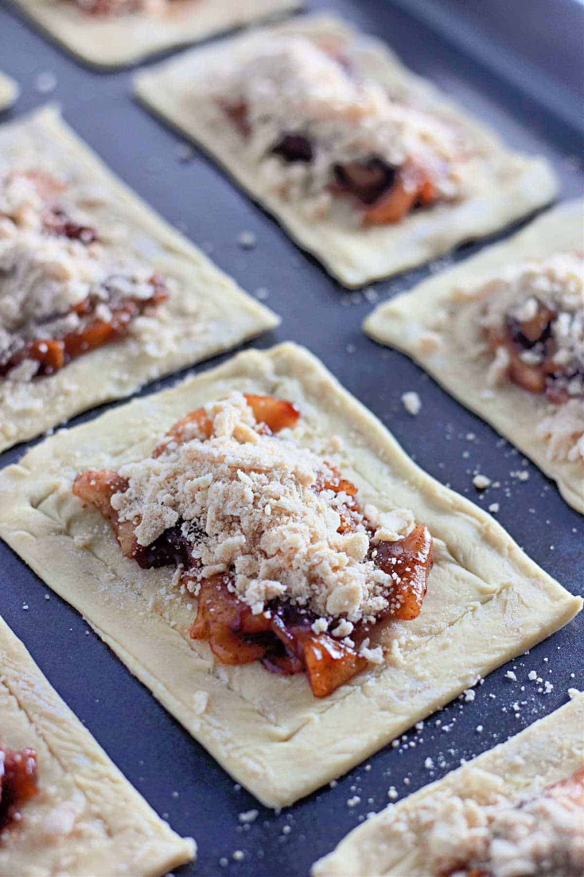 Unbaked rectangular pieces of pastry topped with apple filling and crumb topping on a baking sheet.